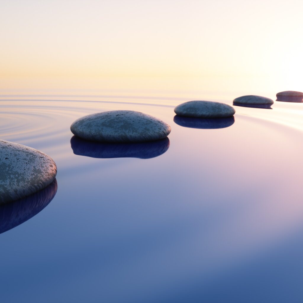 Litteät kivet jonossa tyynessä vedessä, laskeva aurinko heijastuu vedestä.